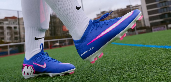 Μπλε και ροζ ποδοσφαιρικά παπούτσια Nike φορεμένα με λευκές κάλτσες σε πράσινο συνθετικό χλοοτάπητα κοντά σε μπλε γραμμή σε εξωτερικό γήπεδο.