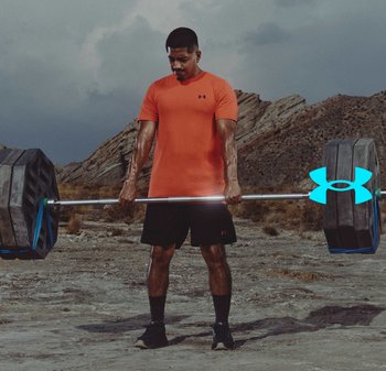 Man in oranje shirt en zwarte korte broek heft een zwaar beladen halterstang buiten op rotsachtig terrein op.