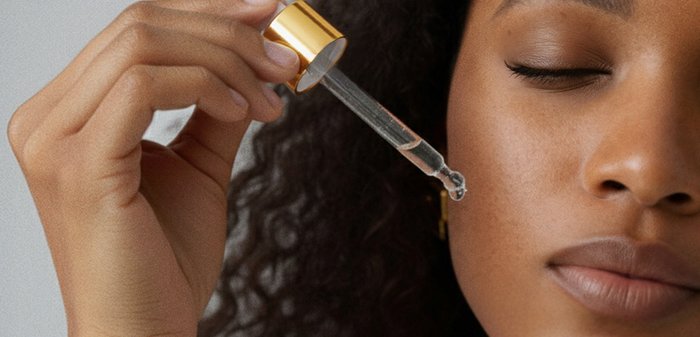 Flacon compte-gouttes avec un capuchon doré tenu au-dessus d'une joue, en train de déposer un sérum clair sur la peau. L'arrière-plan est neutre, mettant en évidence l'application du produit.