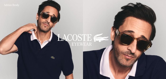 Homme portant des lunettes de soleil foncées et un polo Lacoste bleu marine, posant devant un fond uni gris clair.