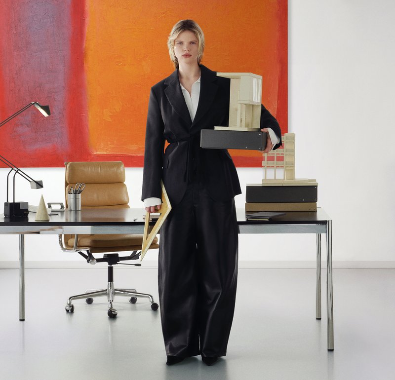 Femme en costume noir tenant des maquettes architecturales et un clipboard, debout dans un bureau moderne avec un bureau, une chaise, une lampe et une peinture abstraite orange.