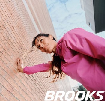 Heldere roze waterdichte jas, met een gestructureerde stof, hoge kraag, ritssluiting, gedragen terwijl er tegen een bakstenen muur geleund wordt.