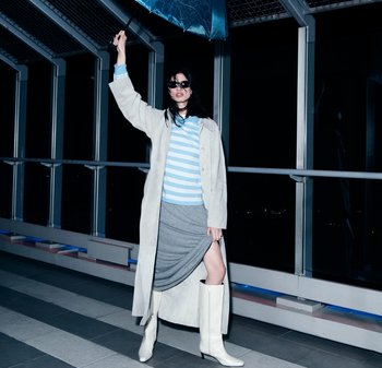 Frau in leichtem Trenchcoat, gestreiftem Hemd, grauem Rock und weißen Stiefeln hält nachts auf einem Dachweg einen blauen Regenschirm hoch.