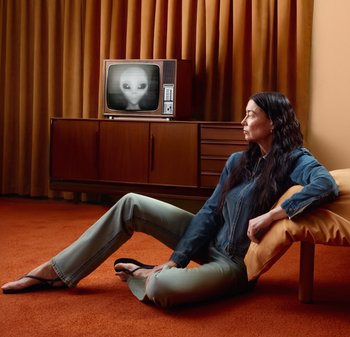 Donna con capelli lunghi e scuri seduta su un tappeto arancione, appoggiata a un cuscino, guarda una TV vintage che mostra un volto alieno in scala di grigi in una stanza retrò.