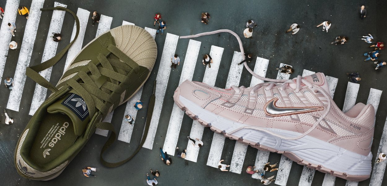 Sneaker Adidas verdi con punta testurizzata e lacci accanto a una scarpa Nike rosa chiaro con tomaia in mesh e dettagli in argento, su un attraversamento pedonale.