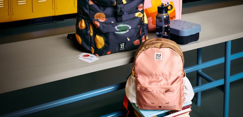 Un sac à dos noir avec des motifs colorés de soleil et de poissons, un sac à dos en forme de chat rose, une boîte à lunch bleue, une bouteille d'eau et des livres sur une table.