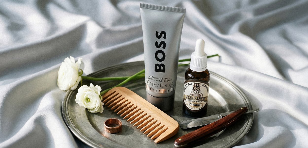 Plateau en métal avec deux fleurs blanches, peigne en bois, bague en cuivre, flacon d'huile à barbe, baume après-rasage BOSS et rasoir droit vintage sur tissu en soie.