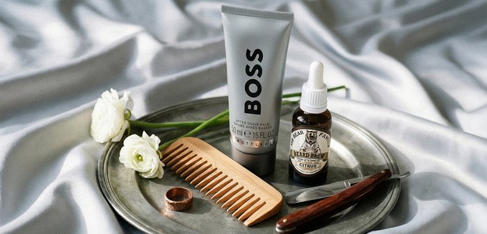 Plateau en métal avec deux fleurs blanches, peigne en bois, bague en cuivre, flacon d'huile à barbe, baume après-rasage BOSS et rasoir droit vintage sur tissu en soie.