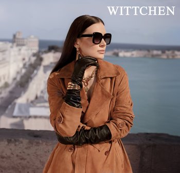 Frau trägt braunen Trenchcoat, schwarze Lederhandschuhe, große schwarze Sonnenbrille und goldenen Schmuck, steht an einem Wasserufer mit einer städtischen Kulisse im Hintergrund.