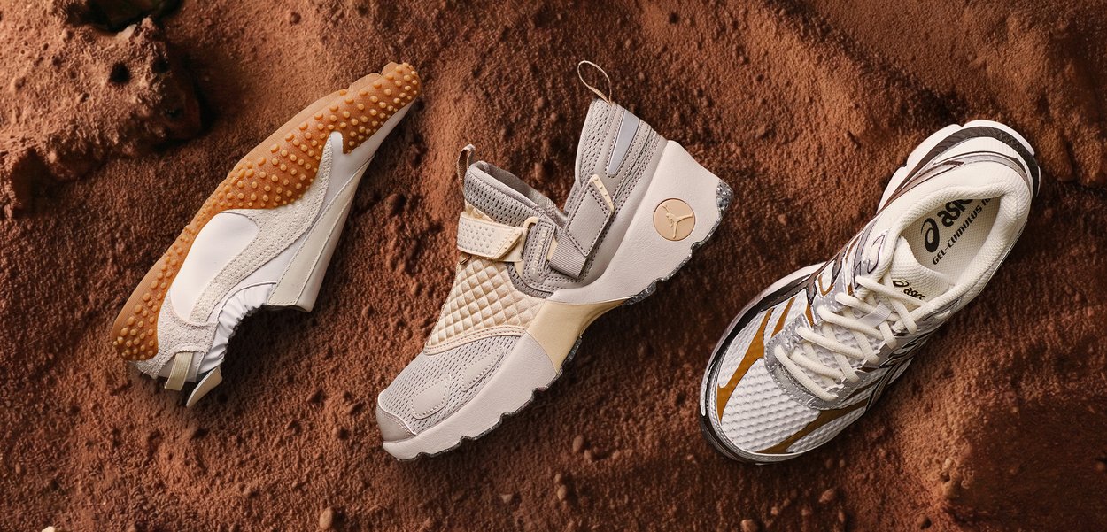 Kolm erinevat kergejõustiku jalanõud beežis, hallis ja valges toonis, paigutatud tekstuurse pruuni muldtausta peale.