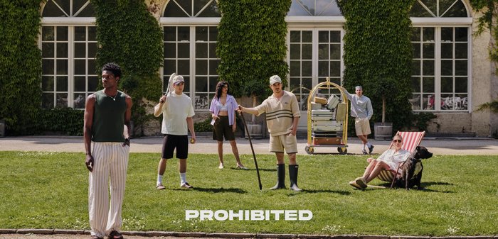 Group of six people in casual summer outfits on green grass. Notable items include a cart with luggage and a dog sitting by a chair.