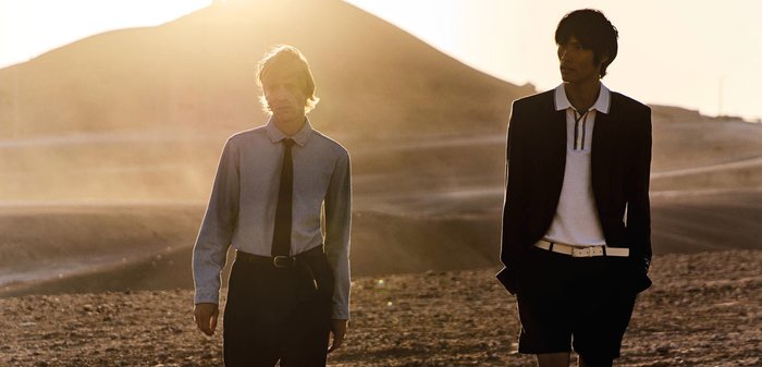 Deux hommes marchent sur un terrain rocheux au coucher du soleil ; l'un porte une chemise claire avec une cravate, l'autre un blazer noir avec un short et une ceinture blanche.