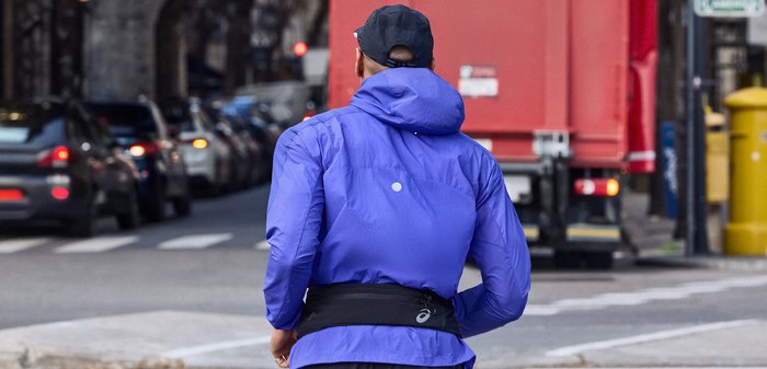 Άτομο με μπλε μπουφάν και μαύρο καπέλο περπατά σε αστική οδό με αυτοκίνητα και κόκκινο φορτηγό στο φόντο κατά τη διάρκεια της ημέρας.