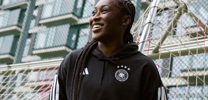 Nasmejana ženska s pletenicami, oblečena v črno športno jopo, stoji pred nogometnim golom na igrišču na prostem.