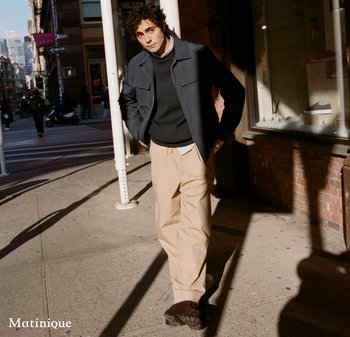 Young man walking on city sidewalk, wearing dark jacket, black sweater, beige pants, and brown shoes, with hands in pockets and sunlight casting shadows.