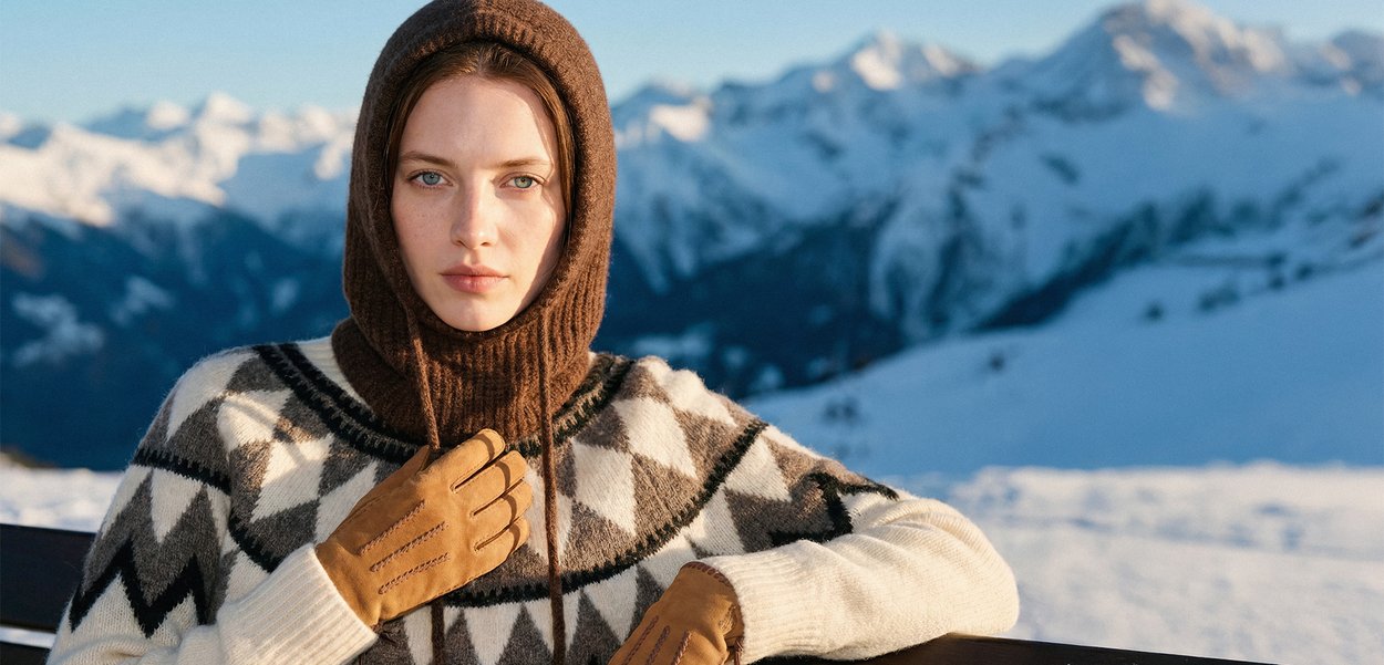 Brūna adīta cepure ar ribotu tekstūru, rakstaina krēmkrāsas un pelēka džemperis, gaiši brūnas cimdi ar šūtām detaļām, sāncens sniegotām kalniem.