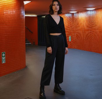 Bluză neagră cropped cu mâneci lungi și sclipici; pantaloni cu dungi subțiri, talie înaltă; bocanci negri cu platformă; decor urban cu plăci portocalii strălucitoare.