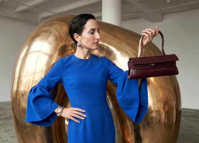 Femme en robe bleue tenant un petit sac à main bordeaux à hauteur des yeux, debout devant une grande sculpture abstraite dorée à l'intérieur.