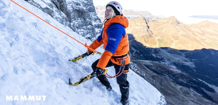 Orangene und blaue puffige Jacke mit strukturierter Oberfläche, Eispickel in der Hand, Klettern auf schneebedecktem Felsen, mit Sicherheitsausrüstung gesichert.