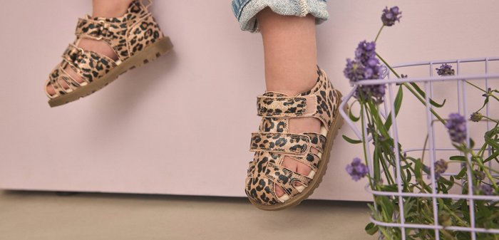 Weiche braune Babyschuhe mit schwarzen Leopardenflecken und einem seitlichen Etikett mit der Aufschrift "EN•FANT", auf einer hellen Stofffläche platziert.