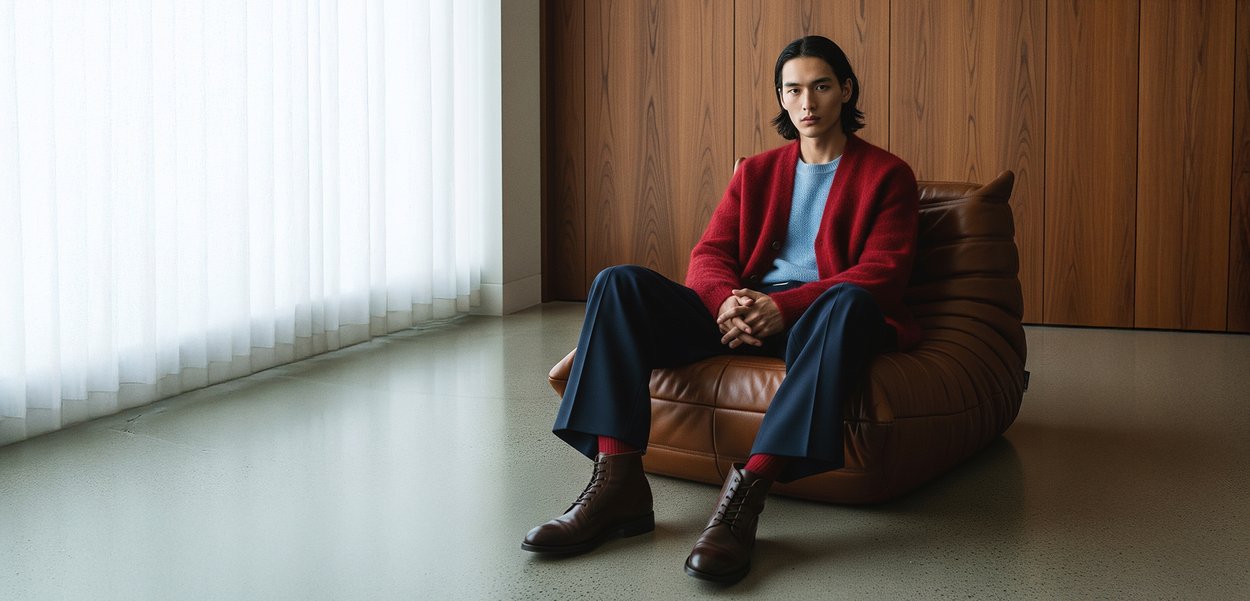 Cardigan rouge sur un pull bleu clair, pantalon bleu marine, chaussettes rouges et bottes marron à lacets. Assis sur un pouf en cuir marron contre un revêtement en bois.
