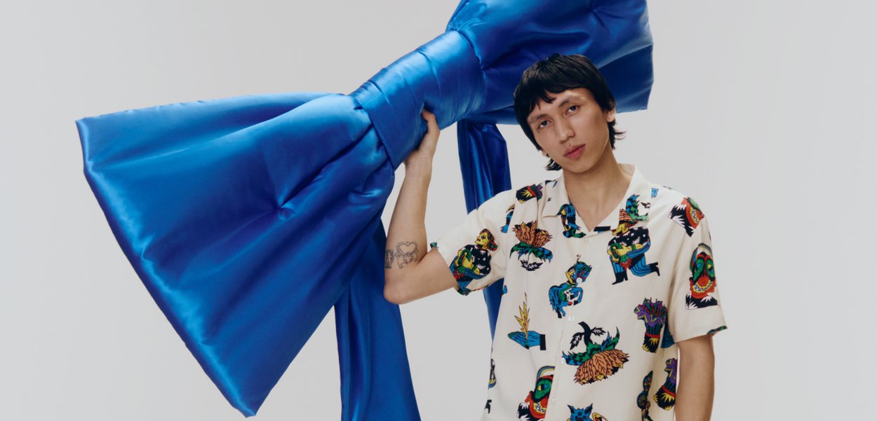 Un grande fiocco blu in satin tenuto sopra la spalla, con una camicia a fantasia che presenta colori vivaci e design astratti.