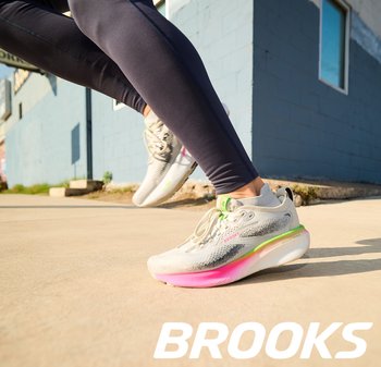 Sportieve schoenen met een lichtgewicht grijze mesh bovenkant, roze en groene accenten, en een gedempte zool, halverwege een stap op het asfalt geplaatst.