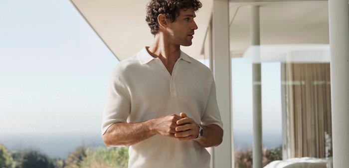 Homme aux cheveux bouclés portant un polo blanc, debout à l'intérieur près de grandes fenêtres, regardant sur le côté les mains jointes.
