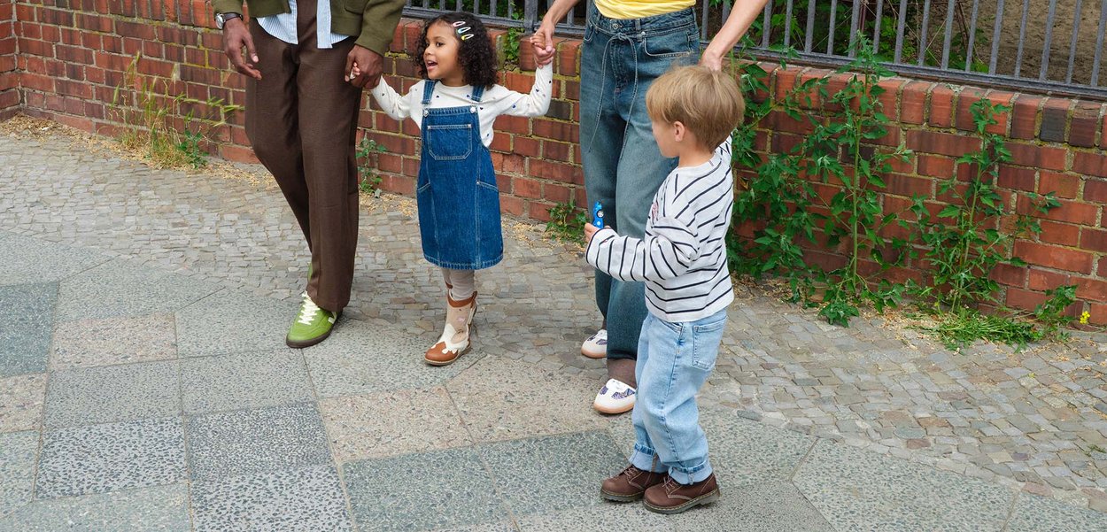 To voksne holder hænder med en smilende pige i en denimkjole, mens en dreng i en striped skjorte står i nærheden på en brostensbelagt fortov.