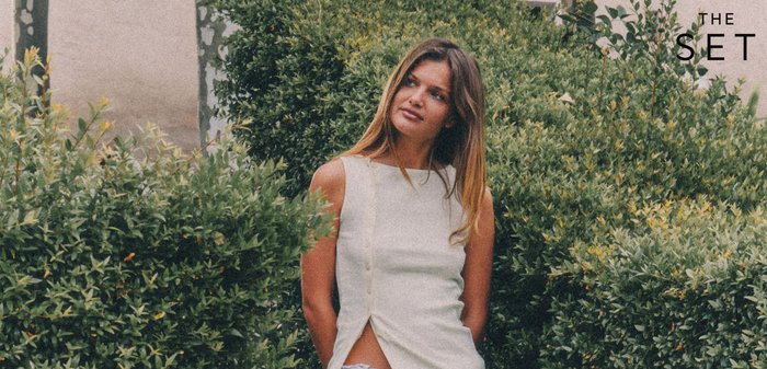 Noor naine valges varrukatuta topis seisab tiheda rohelise põõsaste hulgas, vaadates veidi kõrvale ja nägus rahulik.