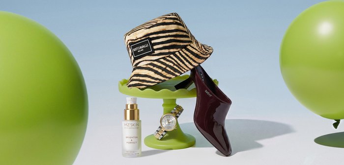 Cappello a secchiello a righe di tigre, scarpa con tacco alto marrone, orologio d’oro su un piedistallo verde e flacone di siero MZ Skin Brighten Perfect tra due palloncini verdi.