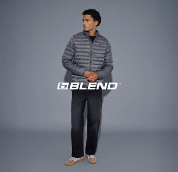 Jonge man die een grijze pufferjas, zwarte broek en beige sneakers draagt, staand tegen een effen grijze achtergrond met het logo "BLEND".