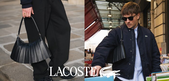 Homme portant des lunettes de soleil et une veste bleu marine traverse la rue en tenant une tasse de café, une baguette et un sac à main noir, parlant au téléphone.
