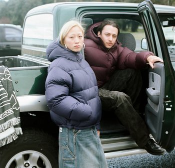 Dos personas están al lado de una camioneta verde. Una lleva una chaqueta de plumas azul marino y una falda de mezclilla; la otra viste una chaqueta de plumas color burdeos y pantalones oscuros.