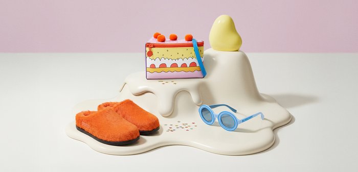 Πορτοκαλί μαλακές παντόφλες, πολύχρωμη τσάντα σε σχήμα τούρτας, μπλε στρογγυλά γυαλιά ηλίου Burberry πάνω σε κρεμ γλυπτή επιφάνεια με μικρά αστέρια κομφετί σε λευκό φόντο.