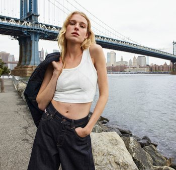 Witte cropped top met een gestructureerd ruitpatroon, gecombineerd met high-waisted donkerdenim broek. De achtergrond toont een brug en een stads-skyline.