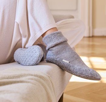 Graue Wollhausschuhe mit strukturiertem gestricktem Bündchen, abgerundeter Zehenpartie und Gummisohle, auf einem hellen Sofa vor einem Holzboden präsentiert.