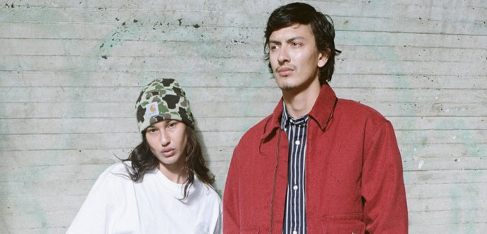 Dos individuos posan frente a un fondo gris texturizado. Uno lleva una camiseta blanca y un gorro verde con estampado, mientras que el otro lleva una chaqueta roja sobre una camiseta de rayas.