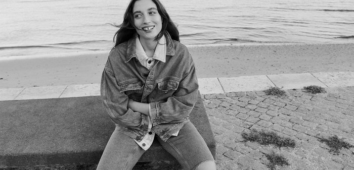 Young woman with long hair wearing a denim jacket sits near a beach with calm water and a horizon in the background.