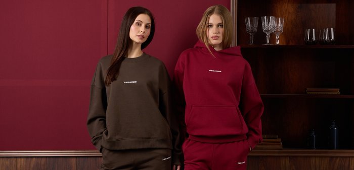 Două femei stau lângă un perete maro cu panouri din lemn, purtând pantaloni de trening și hanorace asortate, de culoare maro și roșu închis.