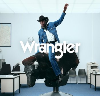 Mann i denimjakke og jeans rir på en mekanisk okse inne på et kontor, med hevet arm for balanse, Wrangler-logo overlagt.