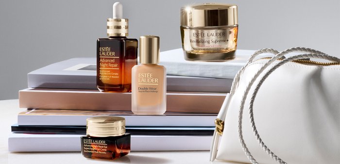 Produits de soins de la peau et de maquillage Estée Lauder disposés à côté d'un sac à main blanc avec des anses en chaînes dorées et argentées.