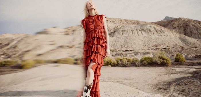 Femme en robe froncée de couleur rouille et bottes en cuir de vache, debout dans un paysage désertique avec des collines rocheuses sous un ciel nuageux.