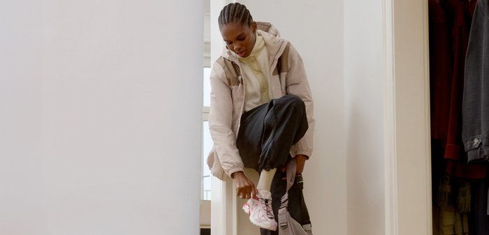Femme portant une veste légère et un pantalon noir, attachant des chaussures de sport blanches et roses en étant assise près d'une porte de placard.