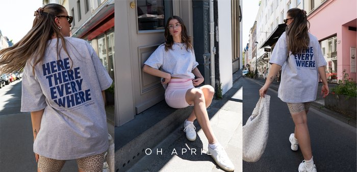 Young woman wearing oversized grey t-shirt with "HERE THERE EVERY WHERE" text and casual sneakers in urban outdoor street settings.