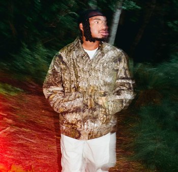 Uomo con dreadlocks che indossa una giacca mimetica e pantaloni bianchi, in piedi all'aperto di notte con effetto di movimento sfocato.