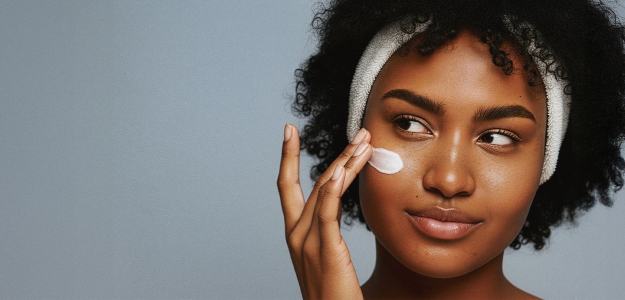 Skincare product application on cheek, using white cream. Model has dark, curly hair, and a white terrycloth headband. Smooth skin texture visible.