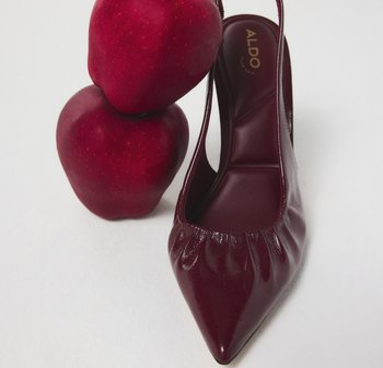 Bordowe lakierowane skórzane szpilki z odkrytymi piętami i spiczastym noskiem, z zebranym wykończeniem na przodzie oraz gładką fakturą, obok dwóch czerwonych jabłek.