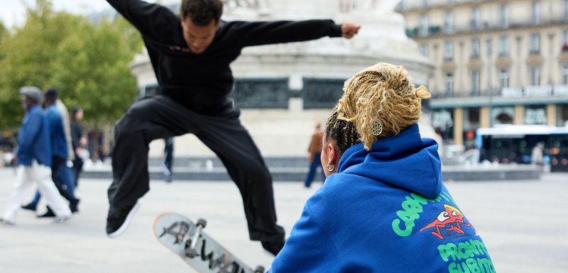 Deux individus dans un environnement urbain. L'un exécute un trick de skate dans une tenue noire ; l'autre porte un hoodie bleu avec des graphismes colorés.