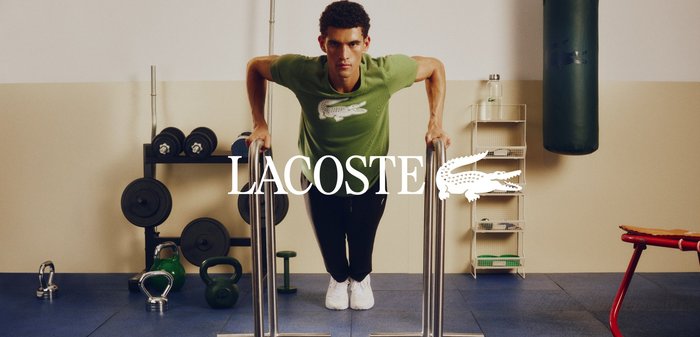 Jeune homme portant un t-shirt et un pantalon Lacoste bleu marine, debout dans une salle de sport tenant une corde à sauter, avec des poids et un sac de frappe en arrière-plan.